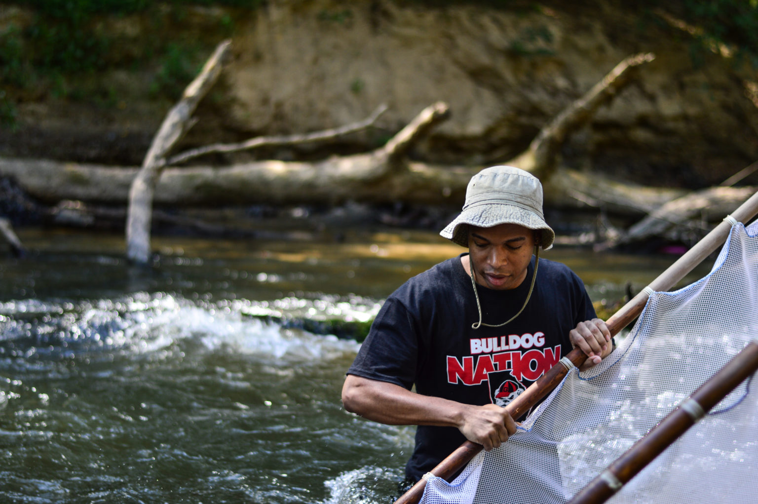 A student wearing a Bulldog Nation t-shirt stands in a stream with a kick-net.
