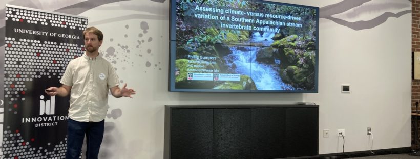Phillip Bumpers presents his research wearing sandals, jeans, and a white collared shirt. A screen displays the image of a stream and text. Above the screen in black and red lettering reads the words Innovation District.