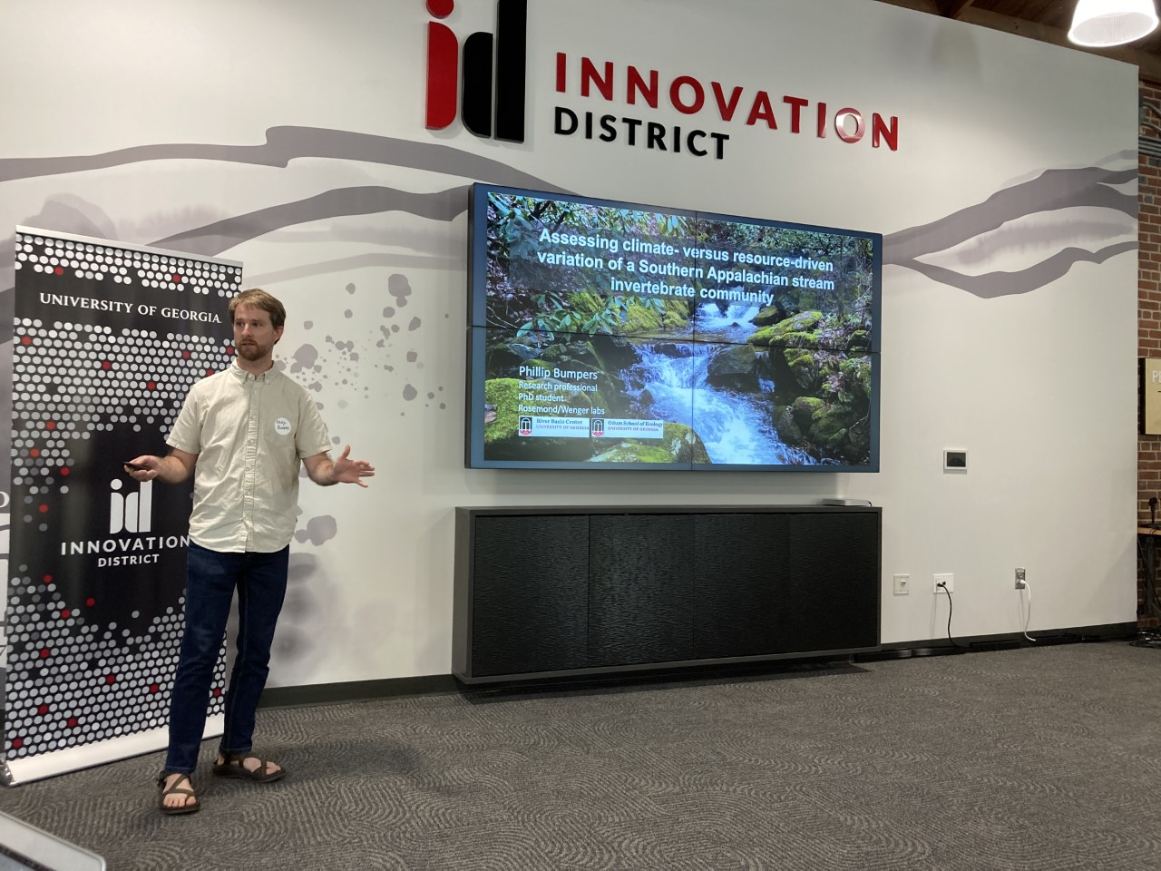 Phillip Bumpers presents his research wearing sandals, jeans, and a white collared shirt. A screen displays the image of a stream and text. Above the screen in black and red lettering reads the words Innovation District.