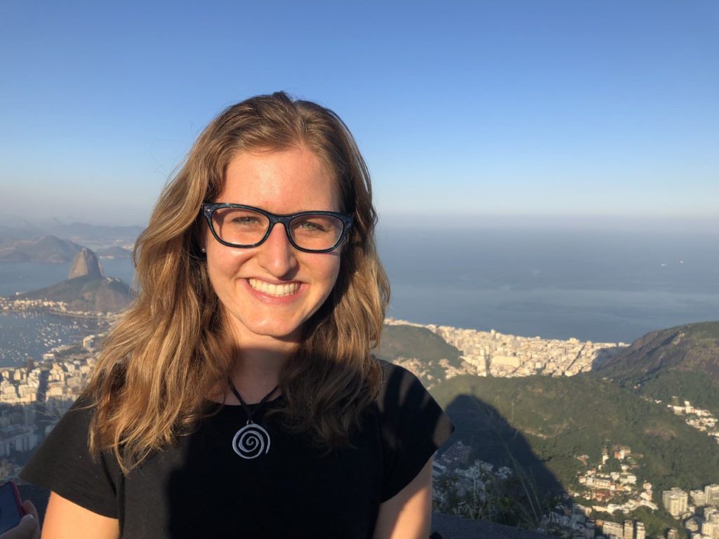 Cydney Seigerman wears black glasses and smiles above a background of hills, buildings and shoreline.