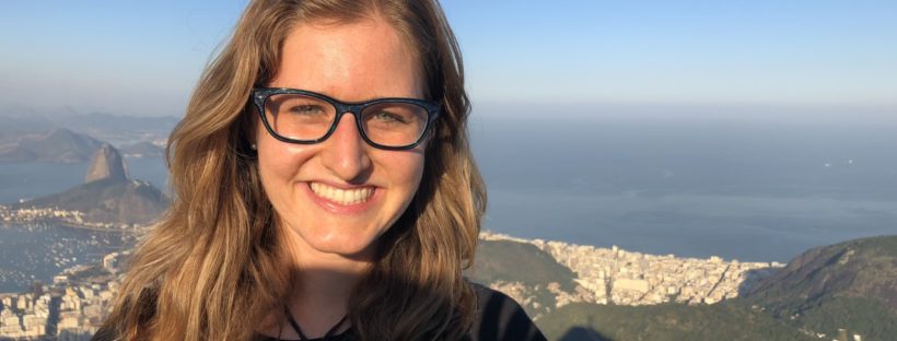 Cydney Seigerman wears black glasses and smiles above a background of hills, buildings and shoreline.