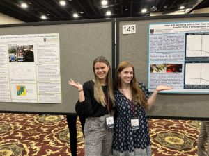 Ally Whiteis, wearing a black blouse and black and white checkered pants, poses with her arm around Olivia Allen, who wears a navy diamond patterned dress in front of their posters.