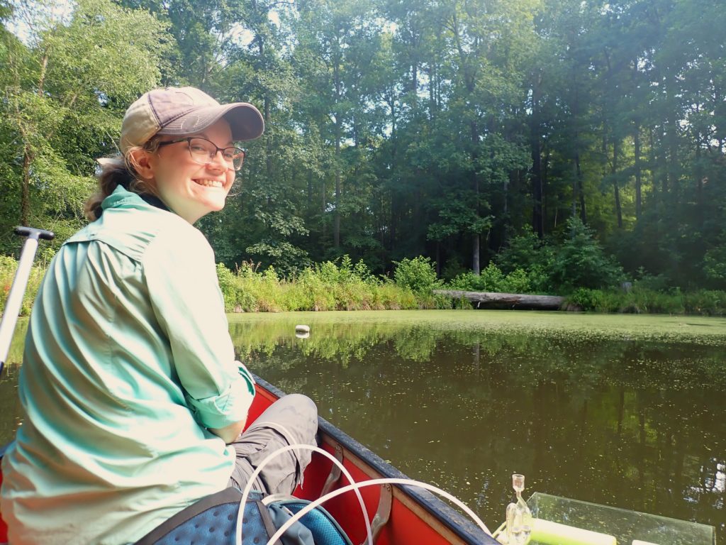 Laura Naslund, sitting in a canoe on a pond, smiles while taking emissions data.