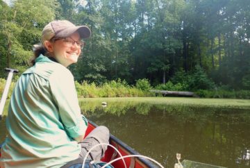 Laura Naslund, sitting in a canoe on a pond, smiles while taking emissions data.