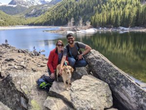 Sechindra Vallury, wearing a backpack, jeans and a gray T-shirt poses with Rebecca Shelton, who wears a red jacket. A light colored dog perches in front of them and behind them Lava Lake in Gallatin Canyon, Montana is visible.