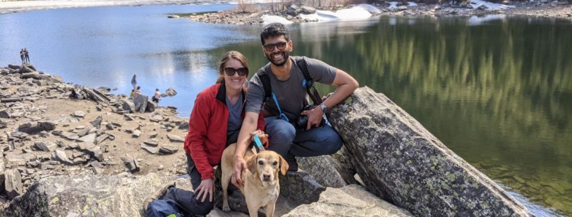 Sechindra Vallury, wearing a backpack, jeans and a gray T-shirt poses with Rebecca Shelton, who wears a red jacket. A light colored dog perches in front of them and behind them Lava Lake in Gallatin Canyon, Montana is visible.