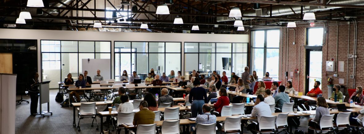A crowd listens to a woman speak, brick wall and exposed ventilation visible in a large room.