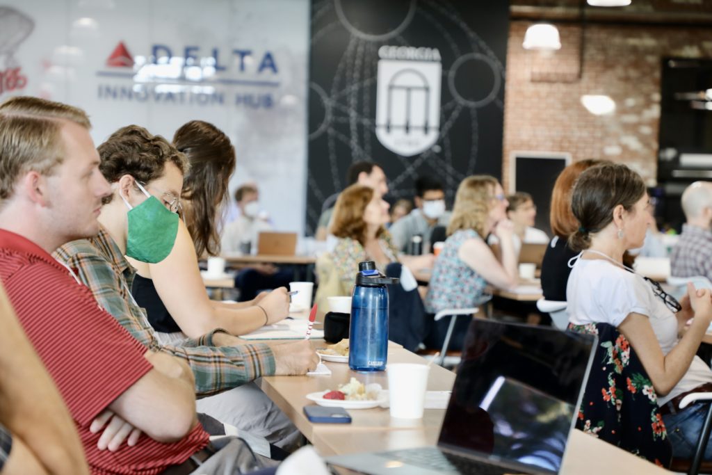 Laptops and water bottles are visible as people listen and take notes.