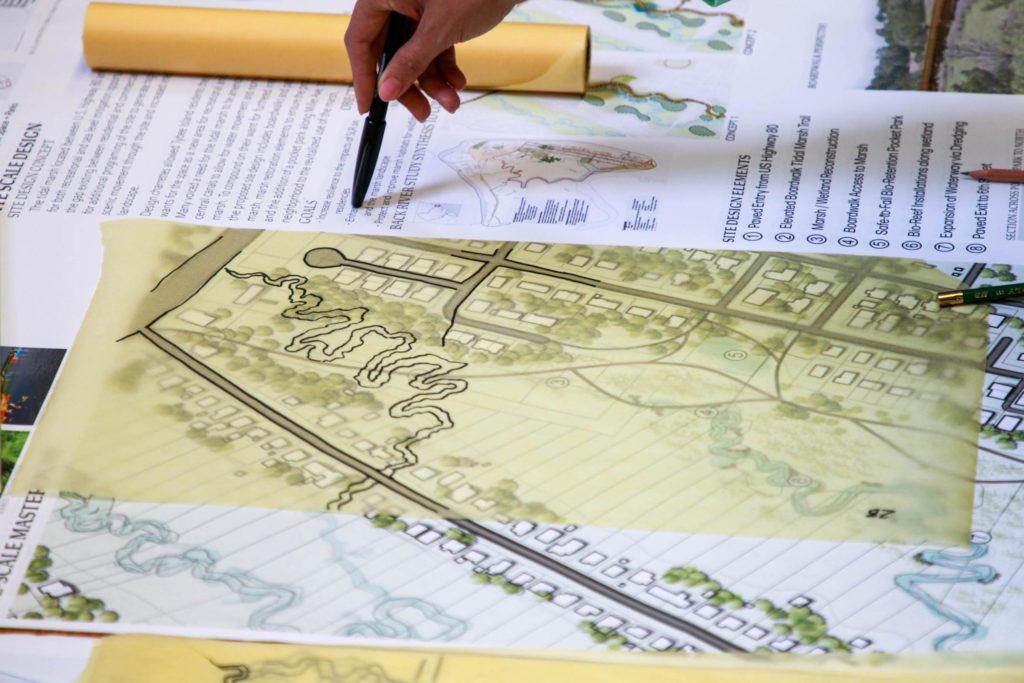 A hand rests the tip of a pen on a large map. Design plans overlay maps of Tybee Island.