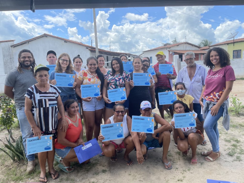 Three rows of people look at the camera, the majority holding up a blue certificate from the cistern informational course. The front row has six people: four people kneeling on the ground, with one person on each side standing up. The second row has six people standing up. The back row has seven people, who are slightly hidden by the second row. In the background, there are two houses with white walls and ceramic tile roofs and one house with a yellow wall.