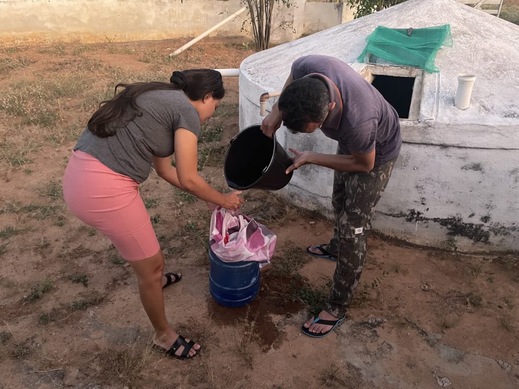 A woman in a pink skirt and gray top secures a pink cloth over a 20L water bottle, as a man in a purple polo shirt and camouflage pants pours water from a black bucket into the bottle. They are in front of a round, white cistern with an angled top, which has a rectangular hole from which water is retrieved from the cistern. The ground is brown dirt with some small patches of grass.