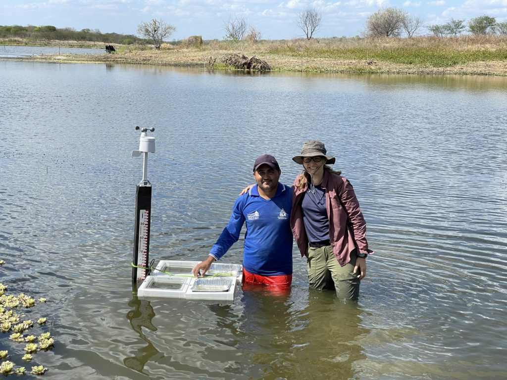 Two people standing together in the blue water of a reservoir, with the water reaching mid-thigh. To the left are four white frames (two with an aluminum foil tray and two with a clear plastic tray) tied together to make a square and secured to a ruler used to measure the water level of the reservoir. On top of the ruler is a white weather station.