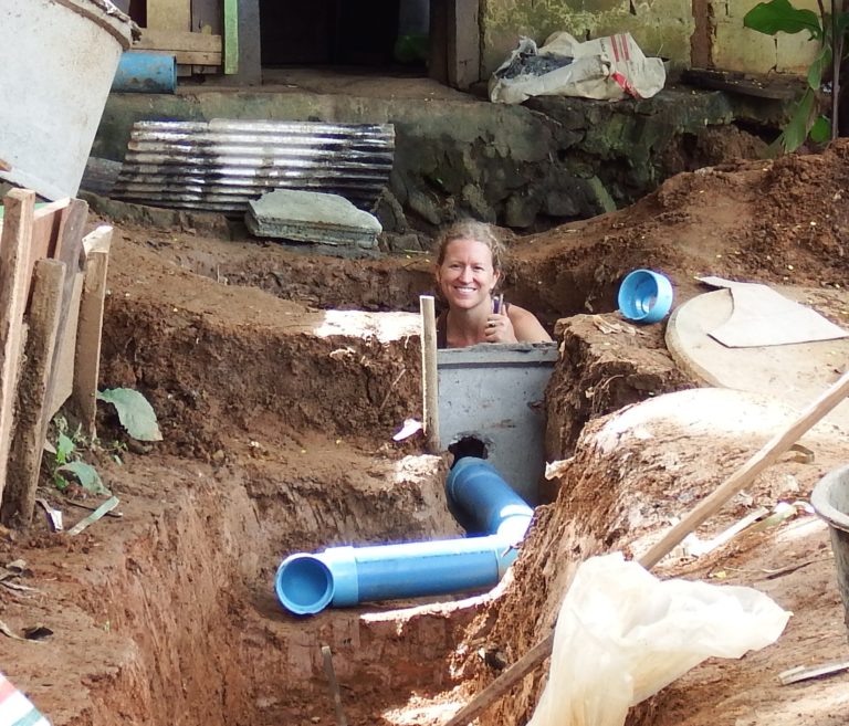 A woman smiles from a dirt pit, pipes and cement visible.