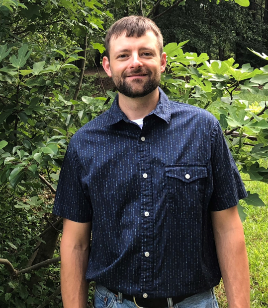 A man in a dark blue button down shirt stands in front of greenery.