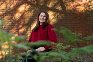Krista Capps stands in front of a brick wall, greenery in the foreground.