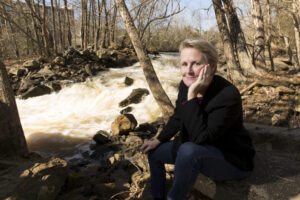 Prof. Emeritus Cathy Pringle sits by a rushing stream, head in her hand, other hand resting on her knee.