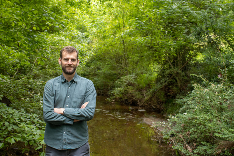 A man stands in front of a stream, arms folded.