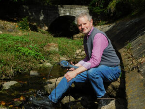 Todd Rasmussen crouches by a river in a vest and purple shirt, holding a blue device.