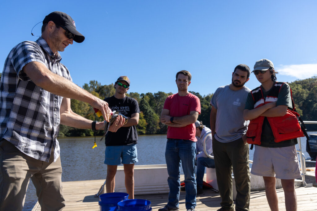 A man on a dock holding research instrument to a fish while students watch.